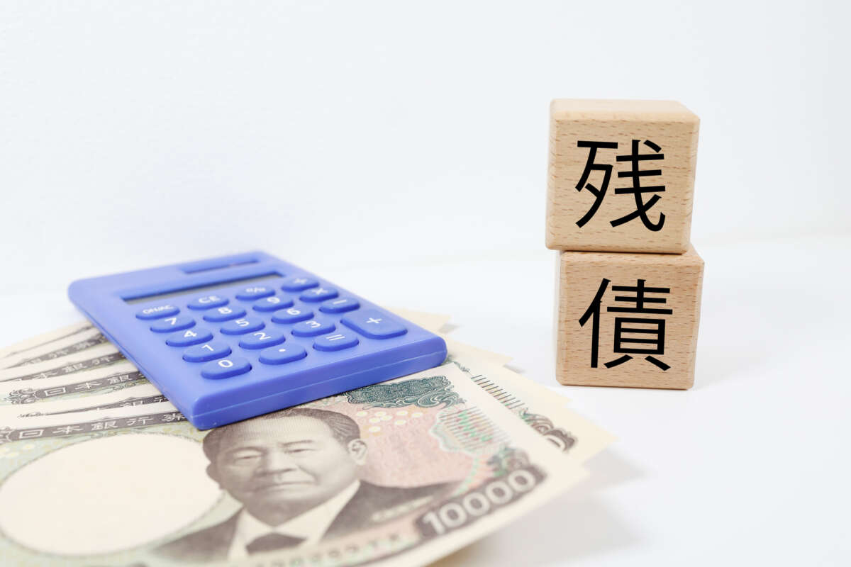 万円札と青い電卓、その横に積まれた「残債」と書かれた木製ブロック