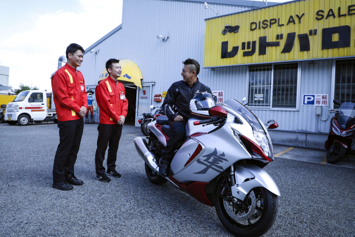 レッドバロンの店舗前でバイクに跨る男性と談笑する2人のスタッフ