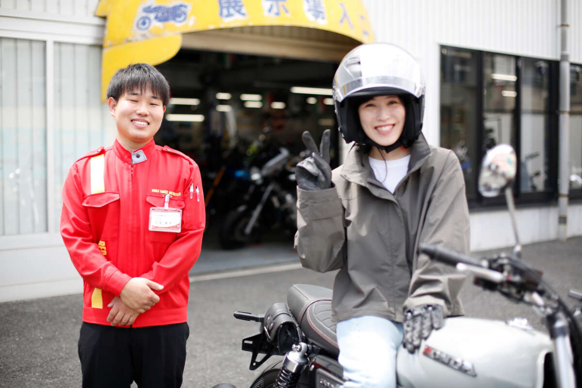 レッドバロンの店舗前でバイクに跨りピースサインをする女性と隣で微笑むスタッフ