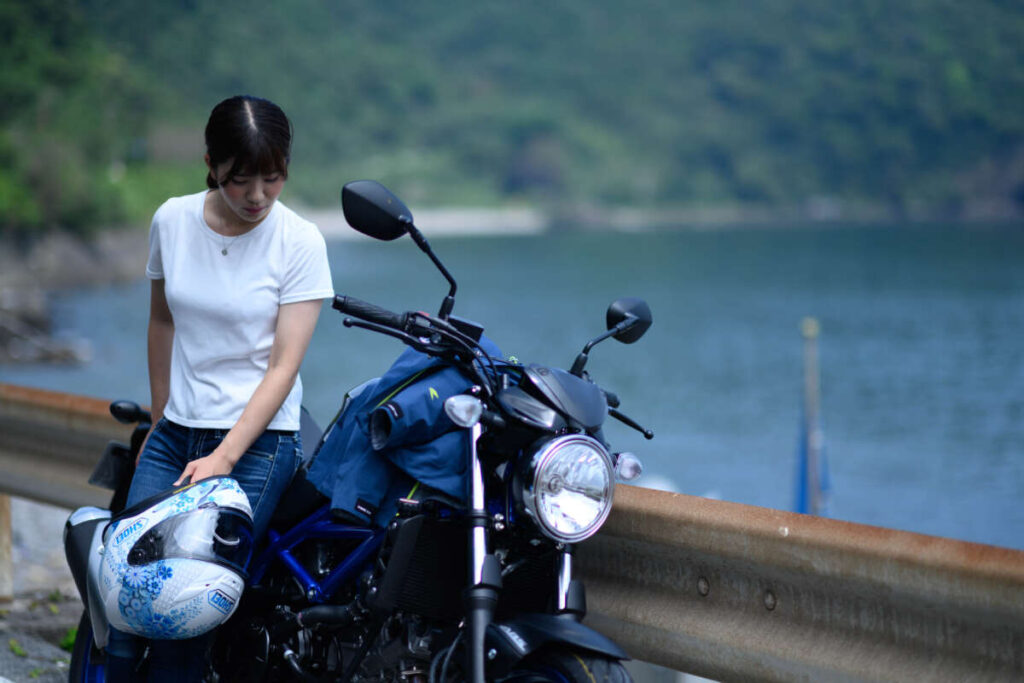 湖畔で女性がバイクのそばに座り、手にしたヘルメットを持ちながら視線を落としている様子