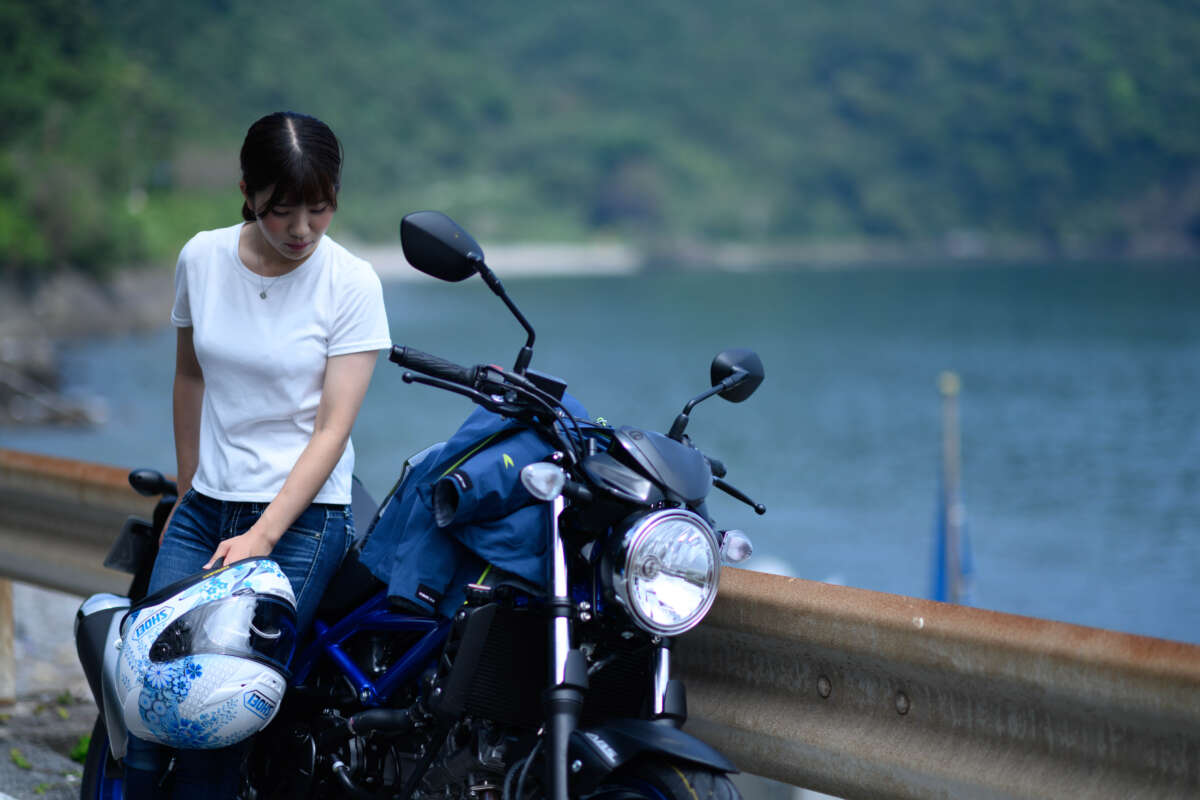 湖畔で女性がバイクのそばに座り、手にしたヘルメットを持ちながら視線を落としている様子