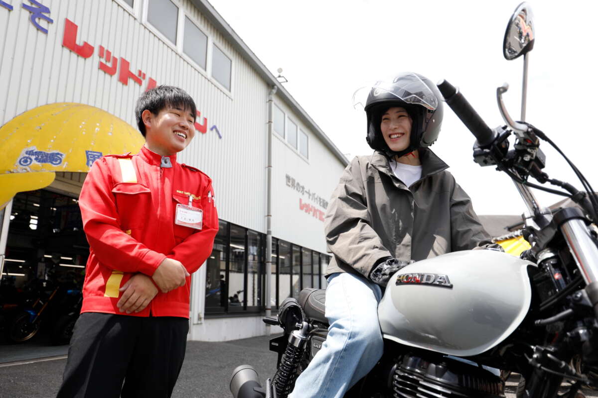 レッドバロンの店先でバイクに跨る女性と笑顔で談笑するスタッフ