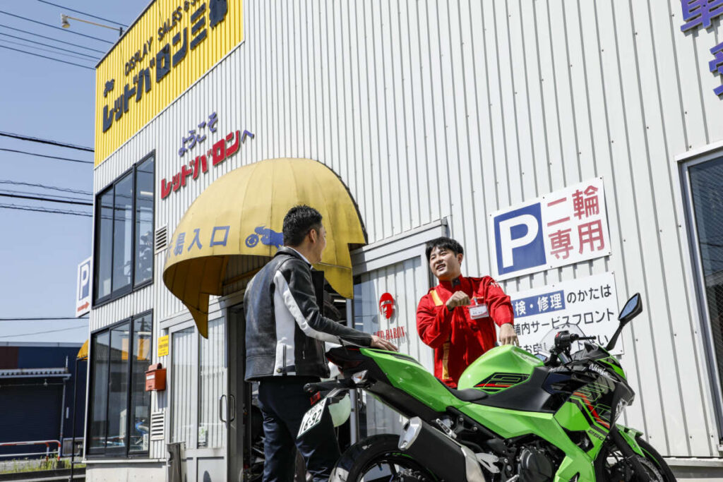 レッドバロンの店舗前で、緑色のバイクを挟んで談笑する従業員と男性
